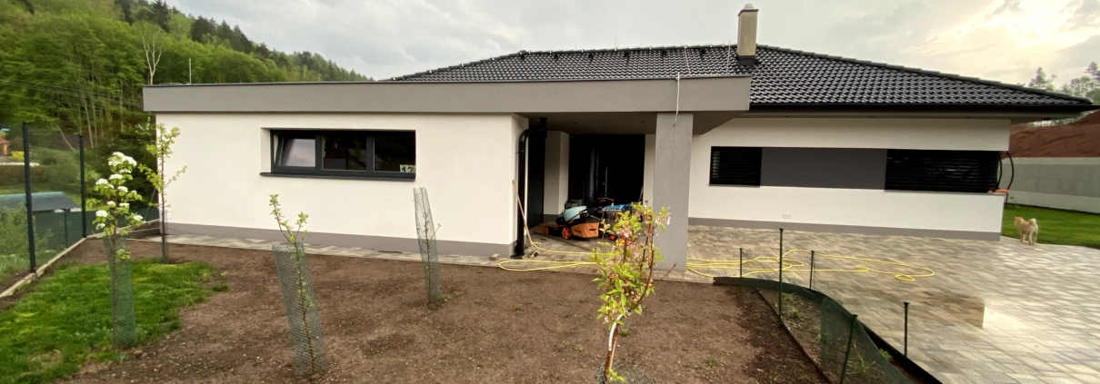Dokončená výstavba bungalov rodinného domu, Garáž v popředí s oknem, Podpěrný sloup, vchodové dveře, dům s okny. Černá střecha.