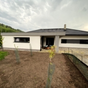 Dokončená výstavba bungalov rodinného domu, Garáž v popředí s oknem, Podpěrný sloup, vchodové dveře, dům s okny. Černá střecha.