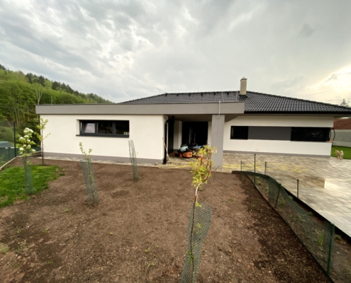 Dokončená výstavba bungalov rodinného domu, Garáž v popředí s oknem, Podpěrný sloup, vchodové dveře, dům s okny. Černá střecha.
